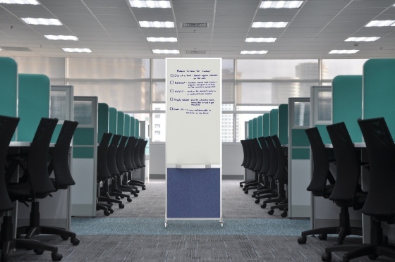 An open-plan office with rows of empty cubicles and chairs facing a central aisle, where a freestanding whiteboard partition stands under bright overhead lights.