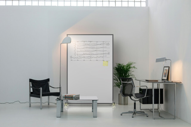 A minimalist office room with a desk, chairs, and lamps, centered around a freestanding white bullet‑resistant whiteboard partition on wheels.