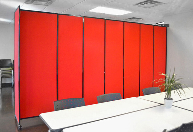  A conference room with tables and chairs in the foreground, backed by a freestanding red bulletproof shield partition made of connected vertical panels.