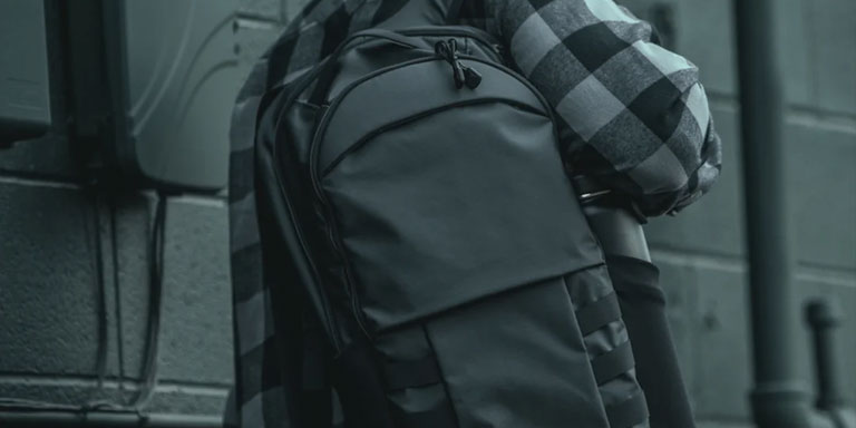 man in gray plaid shirt and gray tactical backpack slung over shoulder walks through urban environment
