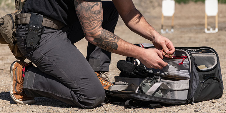 Man at outdoor range kneeling down reaching into range bag with various training items