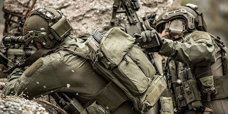 Two military men in full kit. One man reaches for equipment from the other's backpack.'