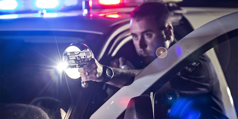 Police officer standing by car with sirens on holding handgun with weapon light illuminated