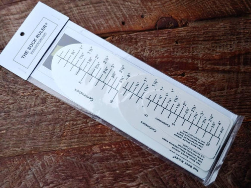 The Sock Ruler, a white ruler with black markings and a grey triangle, for measuring sock sizes on a wooden surface.