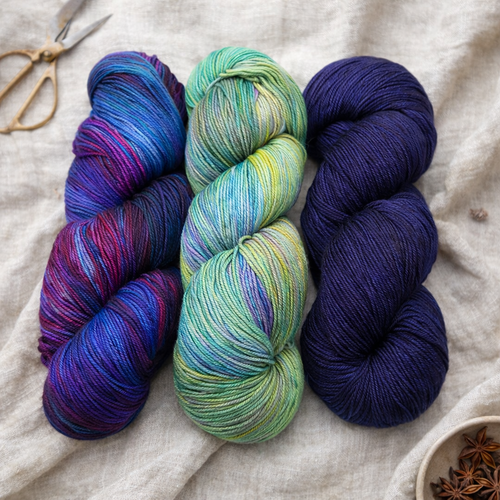 Three skeins of Malabrigo Ultimate Sock Yarn, fingering weight superwash merino nylon, in assorted colors: multicolored purple and blue, green and yellow variegated, and solid dark blue, arranged on a beige fabric with scissors nearby.