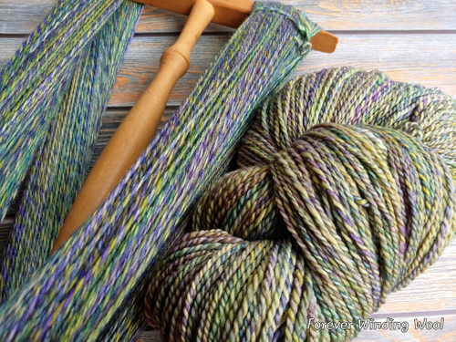 Hand-dyed yarn skein and wooden niddy noddy on a wooden surface, showcasing colorful, multi-colored shades of green, blue, and purple.