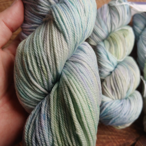Malabrigo Rios yarn skein in Daphne, a soft blend of blue, green, and purple hues. Superwash Merino Worsted yarn, held by a hand, showcasing its gentle color palette and smooth texture, from Malabrigo.