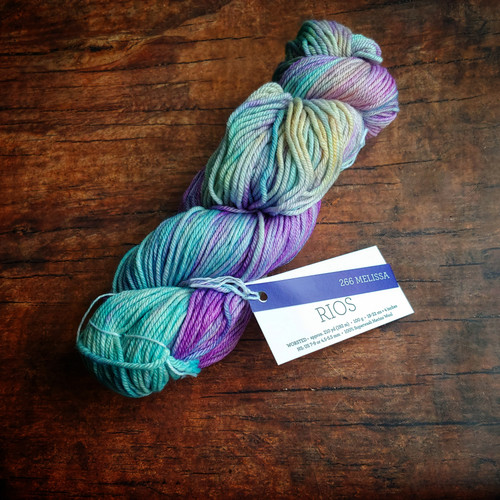 Malabrigo Rios yarn in Melissa color, Superwash Merino Worsted, skein on wooden surface with label. Multicolored yarn featuring shades of purple, blue, green, and yellow hues, neatly wrapped with a band and tag displaying the product name.