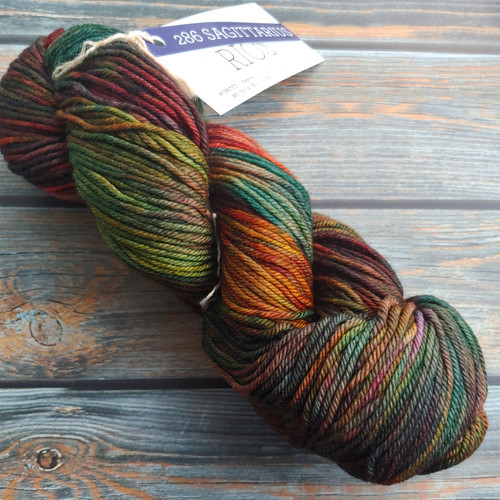 Malabrigo Rios Sagittarius Muted Rainbow Merino Yarn skein with label on wooden surface, showcasing multicolored yarn in shades of green, orange, red, and brown.