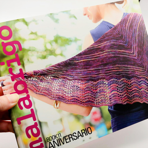 Malabrigo Pattern Book 11: Anniversario, showcasing vibrant multicolored knitted shawl, pink, purple, and blue hues, cover of knitting book.