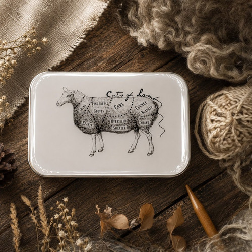 Cuts of Lamb Notions Tin, white metal box with detailed lamb diagram on wooden table with natural fibers and sewing tools nearby.