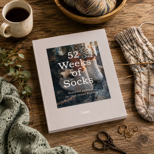 Laine Publishing's 52 Weeks of Socks book cover surrounded by knitting materials, yarn, and a mug on a wooden surface with greenery.
