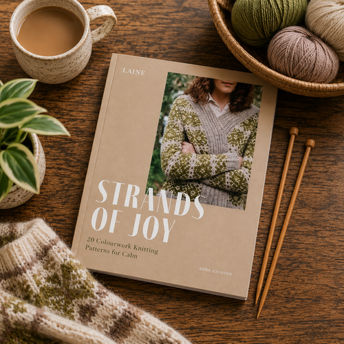Strands of Joy knitting pattern book with 20 colourwork designs, featuring a woman in a green sweater, on a wooden table with yarn and needles.