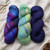 Three skeins of Malabrigo Ultimate Sock Yarn, fingering weight superwash merino nylon, in assorted colors: multicolored purple and blue, green and yellow variegated, and solid dark blue, arranged on a beige fabric with scissors nearby.