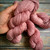 Hand holding mauve pink Hand Dyed Superwash Merino Wool Yarn, showcasing its soft and twisted texture on a wooden background.