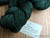 Emerald Green Artisan Merino yarn, Superwash Worsted Weight, dark green skein with subtle sheen, "Forever Winding Wool" label on wooden background, handcrafted wool yarn with luxurious feel and high-quality craftsmanship features.