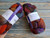 Malabrigo Nube Marte 100% Merino Wool Roving fiber, 4oz, multicolored skeins in purple, orange, and blue hues, suitable for spinning and felting, displayed on a rustic background.