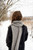 Woman wearing black coat and grey knitted scarf with pattern, standing outdoors in snowy scene, Laine Publishing accessory.