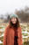 Smiling woman wearing green knitted beanie, red coat and brown sweater, outdoors in snowy field, Laine Publishing 52 Weeks of Accessories inspiration.