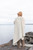 Woman wearing chunky knitwear, beige hat and coat, standing on rocky shoreline overlooking calm water under overcast sky.