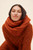 Woman wearing a vibrant burnt orange chunky knit sweater and scarf set, posing against a cream background, exuding cozy elegance and warmth.