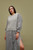 Woman wearing a grey chunky knit sweater and matching striped skirt with a slit, posing against a muted green background.