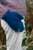 Blue knitted mittens with intricate cable pattern on a person's hand, paired with purple sweater and white pants in a field setting.