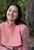 Woman wearing a pink knitted vest and sweater, smiling with eyes closed, posing outdoors in a natural setting with trees in the background.