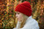 Red knit beanie on a woman with blonde hair, wearing a white sweater, surrounded by autumn foliage with yellow and orange leaves.