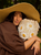 Woman in brown dress and straw hat holding white crocheted bag with daisies, smiling outdoors in a natural setting.