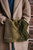 Green and brown knitted scarf with pattern, tucked into a tan coat, displayed against a rustic wooden background, 52 Weeks of Scarves.