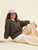 Woman wearing a purple hat, brown sweater, and beige pants sitting on the floor against a beige background, showcasing casual knitwear style.
