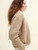 Woman wearing beige knitted sweater with textured pattern, paired with white pants, showcasing side profile against a cream background, Laine Publishing knitting inspiration.