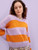 Woman wearing lilac and orange striped knitted sweater, floral patterned bottoms, standing against light purple background, showcasing Laine Publishing style.
