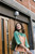 Woman wearing green and peach knitted top, standing in front of wooden doors with a building number sign, Laine Publishing knitting inspiration.