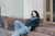 Woman with long brown hair wearing blue jeans and dark green sweater sitting on stone steps outside building with wooden door and window.