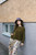 Woman wearing green sweater, floral pants, and blue hat poses against building with window, showcasing 52 Weeks of Easy Knits beautiful patterns.
