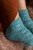 Teal knitted socks with intricate patterns on a person's feet, paired with rust-colored clothing, showcasing beautiful designs from 52 Weeks of Socks, Vol. II.