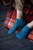 Woman's feet wearing blue knitted socks with intricate lace patterns, resting on red plaid blanket beside a wooden chair on a rug.