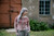 Woman wearing a pink knitted cardigan with colourwork pattern, standing in front of a rustic building, showcasing Strands of Joy knitting design.
