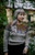 Woman wearing grey, orange, and blue knitted sweater with intricate colourwork design, standing outdoors beside greenery with a building in background.