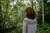 Woman with long brown hair wearing grey sweater with intricate dark grey colourwork pattern on shoulders, standing in a forest with green trees.