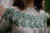 Woman wearing grey sweater with green floral colourwork pattern, showcasing Strands of Joy knitting design on the chest and sleeves.