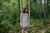 Woman wearing grey knitted dress with blue pattern, standing in forest, showcasing Strands of Joy colourwork design, calm atmosphere.