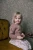 Blonde girl sitting on mustard chair wearing pink sweater and floral skirt holding black and white beads with green and beige wallpaper background.