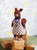 Brown knitted squirrel toy wearing white and red checkered dress and blue necklace, standing on stacked boxes, from Mouche & Friends by Laine Publishing.