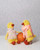 Two yellow knitted duck toys, one in pink and white scarf and dress, other in multicolor dress, standing with orange ball, by Laine Publishing.
