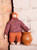 Mouche & Friends seamless knit monkey toy wearing purple sweater and orange pants standing beside a brown clay pot on a stone shelf.