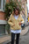 Woman wearing a yellow knitted cardigan, white collared shirt, and black pants standing on a street with buildings and flowers in the background.