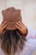 Woman wearing brown knit beanie and white sweater, adjusting hat with hands adorned with rings, showcasing casual winter fashion and effortless style.